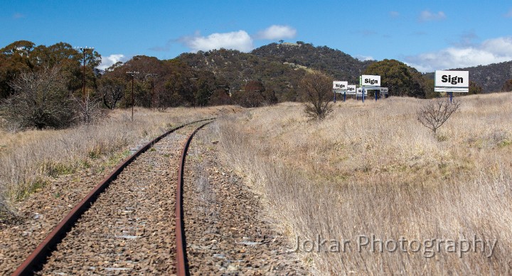 Railway.jpg - Railway (Sign Sign Sign Sign Sign Sign).  Even before the railway reached Goulburn in 1869, there were calls for the line to be extended to the Monaro. The line to Cooma (through Queanbeyan, Michelago, Bredbo and Bunyan) was eventually opened in 1889, with later extensions through to Nimmitabel (1911) and Bombala (1921).The arrival of the railway had a huge social and economic impact, and really served to ‘open up’ the region to the outside world. However, despite a boom during the construction of the Snowy Scheme, when Cooma was the base of operations, the line always ran at a loss. One justification for the railway had been that it would allow for shipment of wheat grown in the Monaro to other markets - but it had quite the opposite effect. The marginal Monaro grain was unable to compete with the cheaper wheat that could now be shipped in from other regions.The line closed in 1988 - exactly one hundred years after its establishment. The billboard signs now have no rail passengers to talk to (though they still ‘serve’ the adjacent highway traffic). We tend to look at billboards in terms of the messages they display, rather than as large constructions plonked onto the landscape. They (almost) never draw attention to themselves as objects. We are so accustomed to their presence as we travel that we don’t really take notice of their physicality either.But these signs, photographed along the now-closed Cooma rail line, have nothing to say about fast food outlets, ski resorts or car repairs. Their only purpose is to declare their own ‘sign-ness’. 